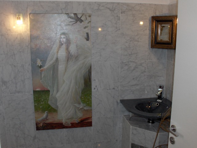 Łazienka wykończona marmurowymi płytkami z umywalką narożną i obrazem kobiety w bieli, oświetlona kinkietem, z lustrem w ozdobnej ramie.